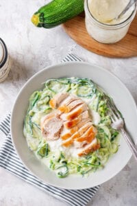 A bowl of zuchinni noodles that have been covered in alfredo sauce, and topped with a cut-up chicken breast.