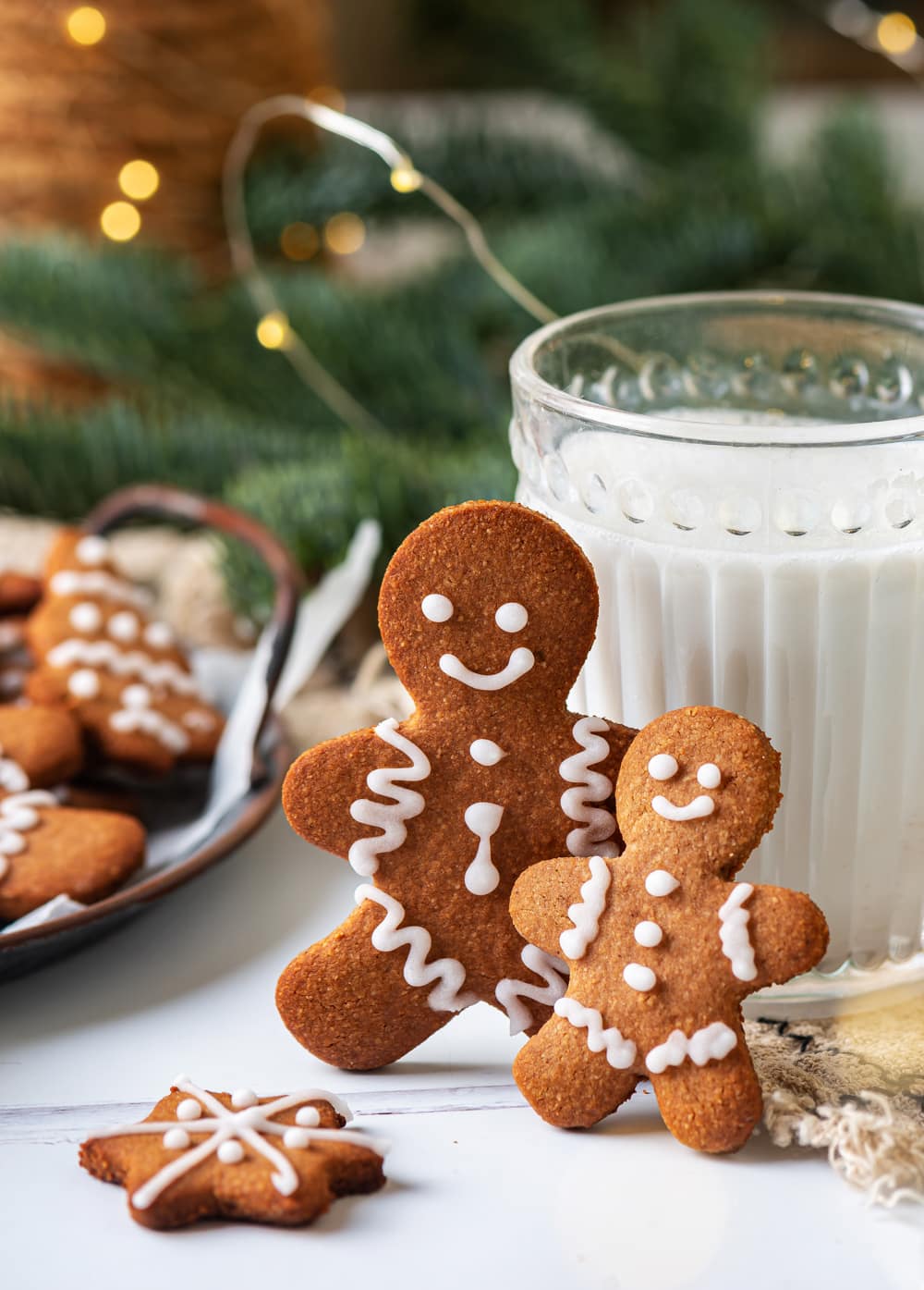 Two gingerbread cookies that are leaned against a glass of milk, with green pine branches and gold lights behind them.