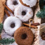 Four glazed gingerbread donuts and one plain gingerbread donut scattered on top of a piece of paper. Two sticks of cinnamon are surrounding the donuts on each side. Pine branches are at the bottom left and top right of the paper.