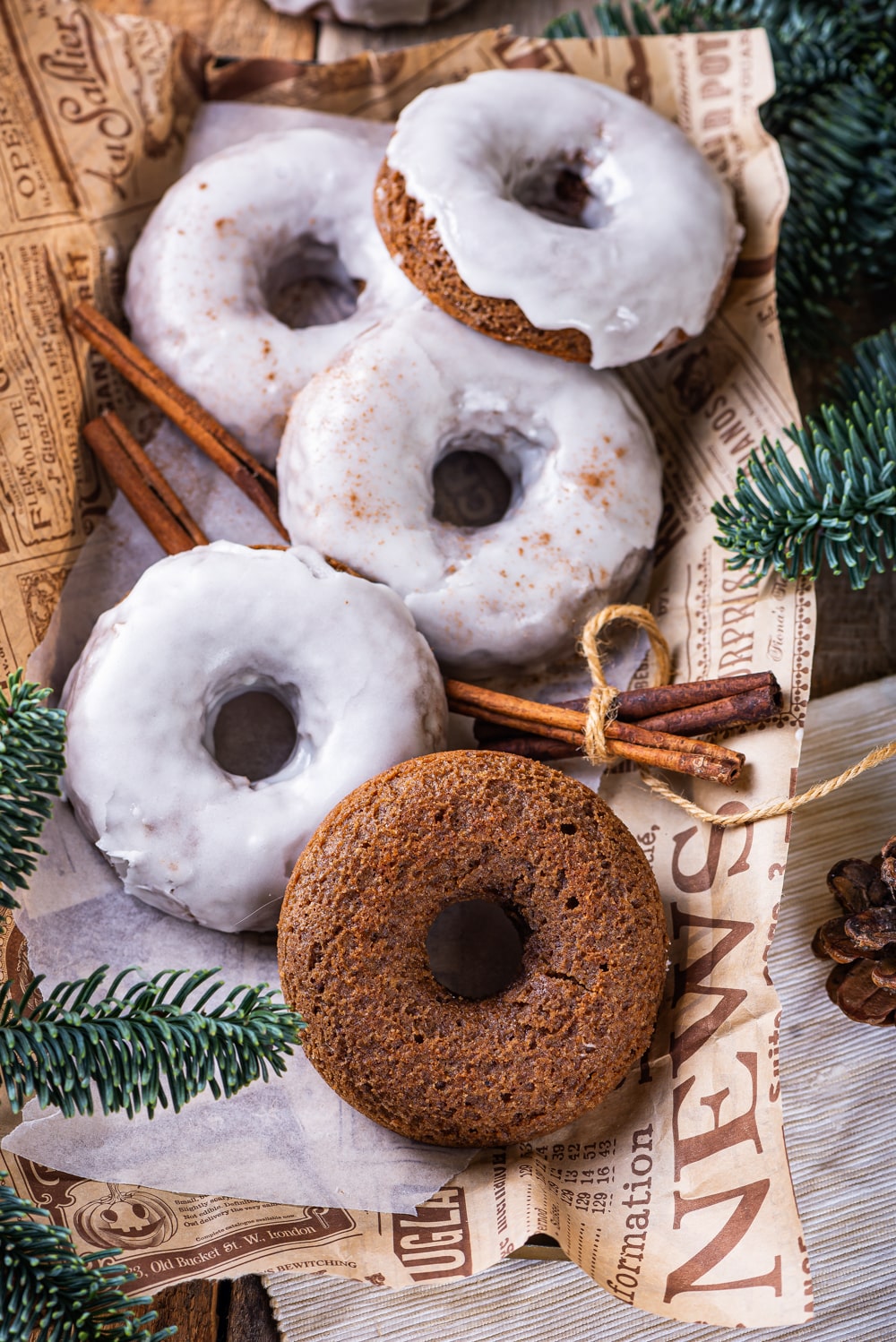 Four glazed gingerbread donuts and one plain gingerbread donut scattered on top of a piece of paper. Two sticks of cinnamon are surrounding the donuts on each side. Pine branches are at the bottom left and top right of the paper.