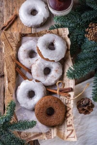 Five glazed gingerbread donuts and one plain gingerbread donut scattered on a piece of paper with two sticks of cinnamon on the sides of the donuts. Pine branches are at the bottom left and top right of the paper. One glazed gingerbread donut is set behind the piece of paper. Everything is on top of a wooden table.