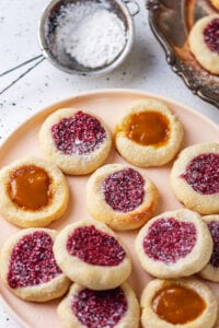 Thumbprint cookies on a plate. Some of the cookies are filled with raspberry jam, and some are filled with peach jam.