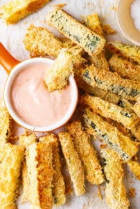 A zucchini fry dunked inside of a small cup of dipping sauce with other zucchini fries around it.