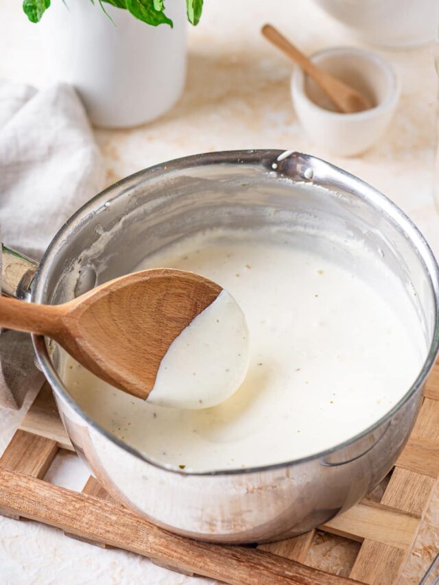 A stainless steel bowl filled with alfredo sauce with a wooden spoon hovering above the alfredo sauce with sauce on the front half of the head of the spoon.