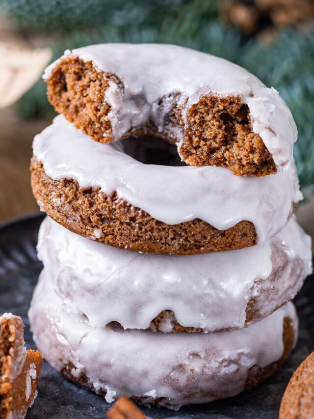 A stack of four glazed gingerbread donuts on a plate. The donut at the top of the stack is cut in half.