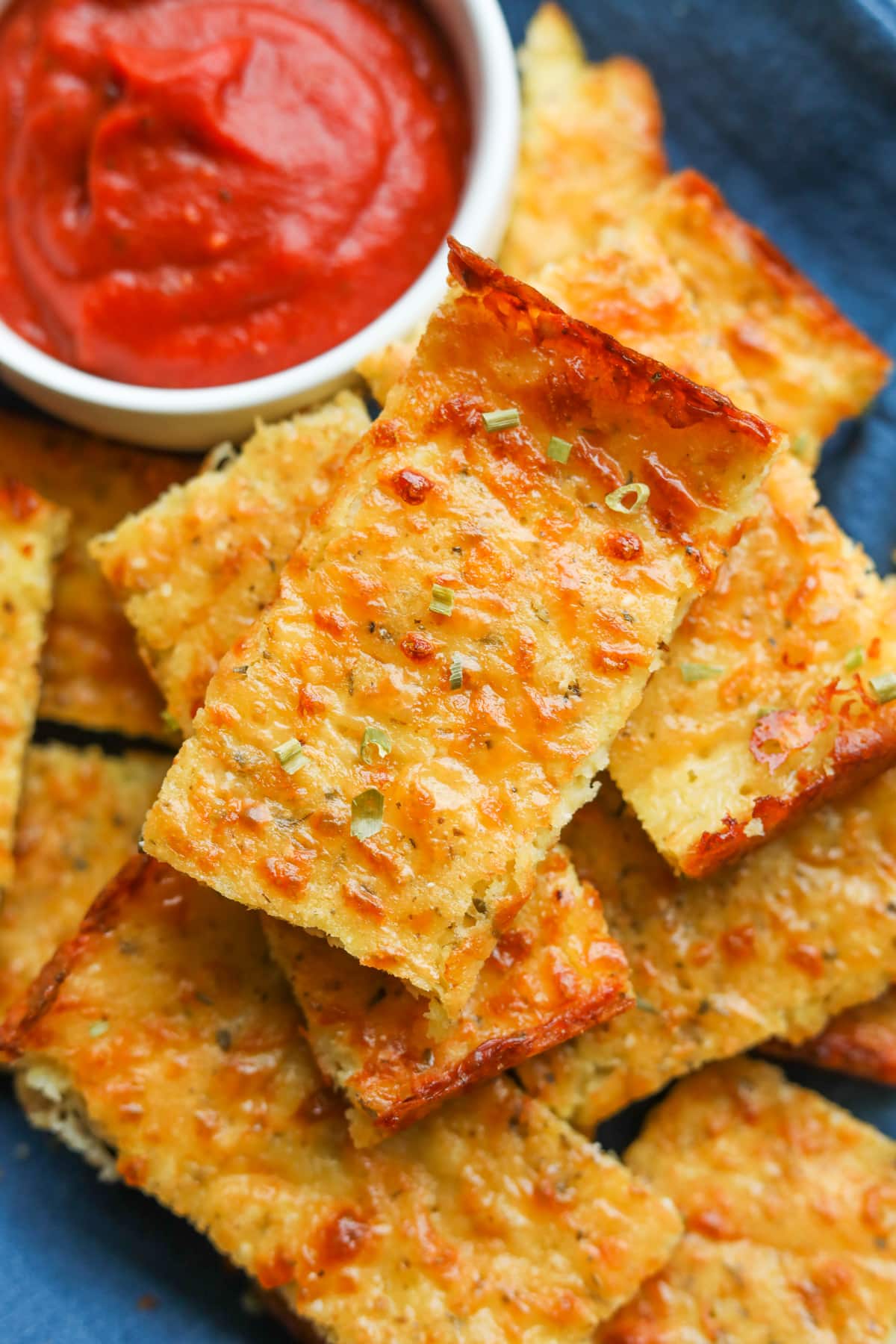 Slices of cheese bread on a blue napkin. The cheese bread is topped with green onions and there is a white cup with marinara sauce in it on the napkin as well.