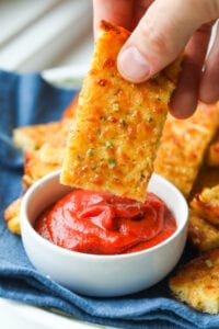 A hand holding a slice of cheese bread over a cup of marinara sauce.