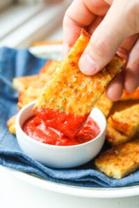 A hand holding a piece of cheese bread that has marinara sauce on the end of it. There is a white cup filled marinara sauce below the piece of bread.