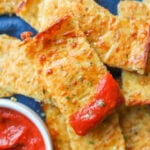 Pieces of cheese bread stacked on top of each other on a blue napkin. The top piece of cheese bread has marinara sauce on the end of it, and there is a cup of marinara sauce next to it.