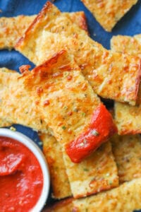 Pieces of cheese bread stacked on top of each other on a blue napkin. The top piece of cheese bread has marinara sauce on the end of it, and there is a cup of marinara sauce next to it.