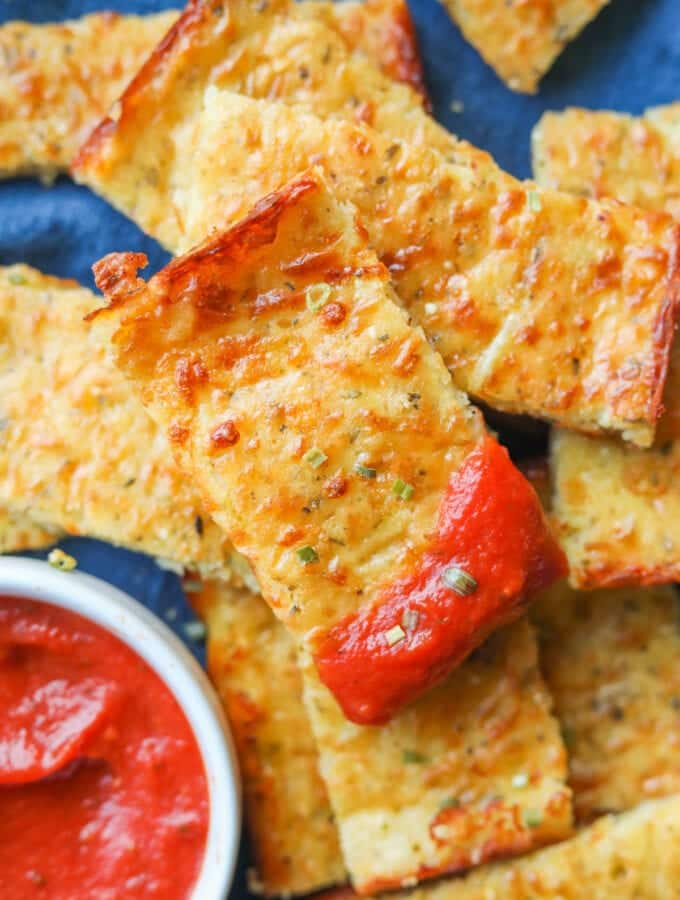 Pieces of cheese bread stacked on top of each other on a blue napkin. The top piece of cheese bread has marinara sauce on the end of it, and there is a cup of marinara sauce next to it.