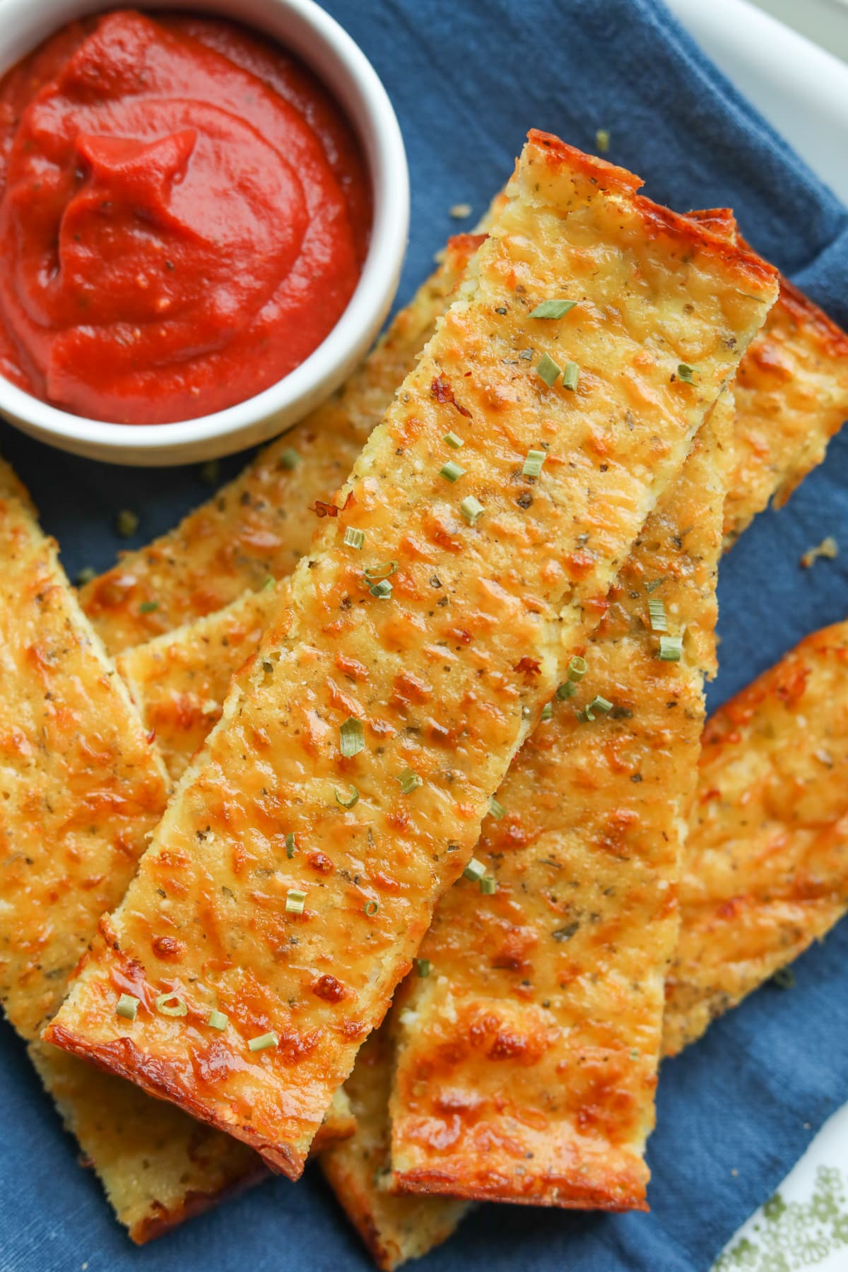 Strips of cheese bread piled on top of one another on a blue napkin. A small white cup of filled with marinara sauce is next to the pieces.