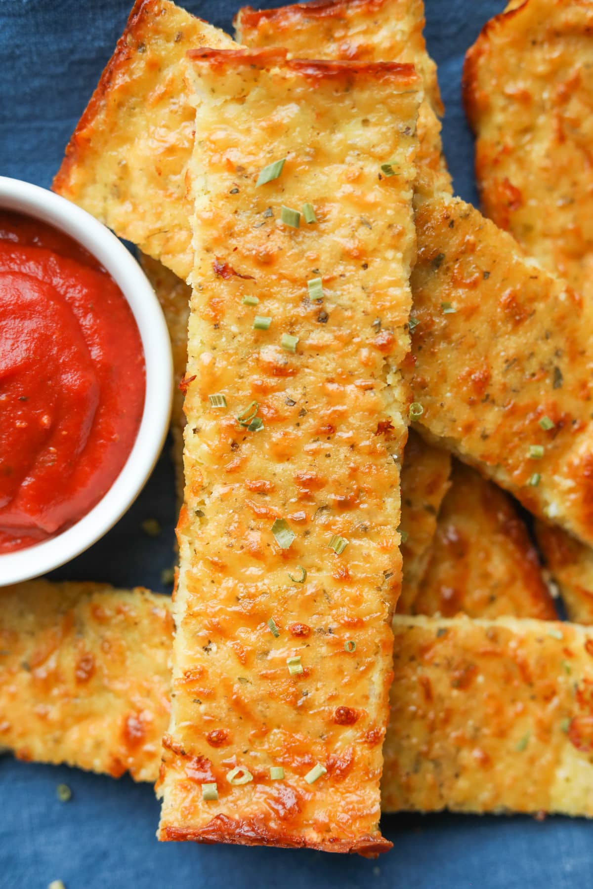 Slices of cheese bread stacked on top of a blue napkin with a cup of marinara sauce next to them.