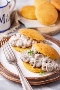 2 Biscuit and gravy sandwiches on a plate, with more biscuits behind them on a cutting board.