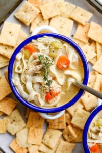 A bowl of chicken noodle soup that's surrounded by cheese crackers. There's a spoon in the bowl.