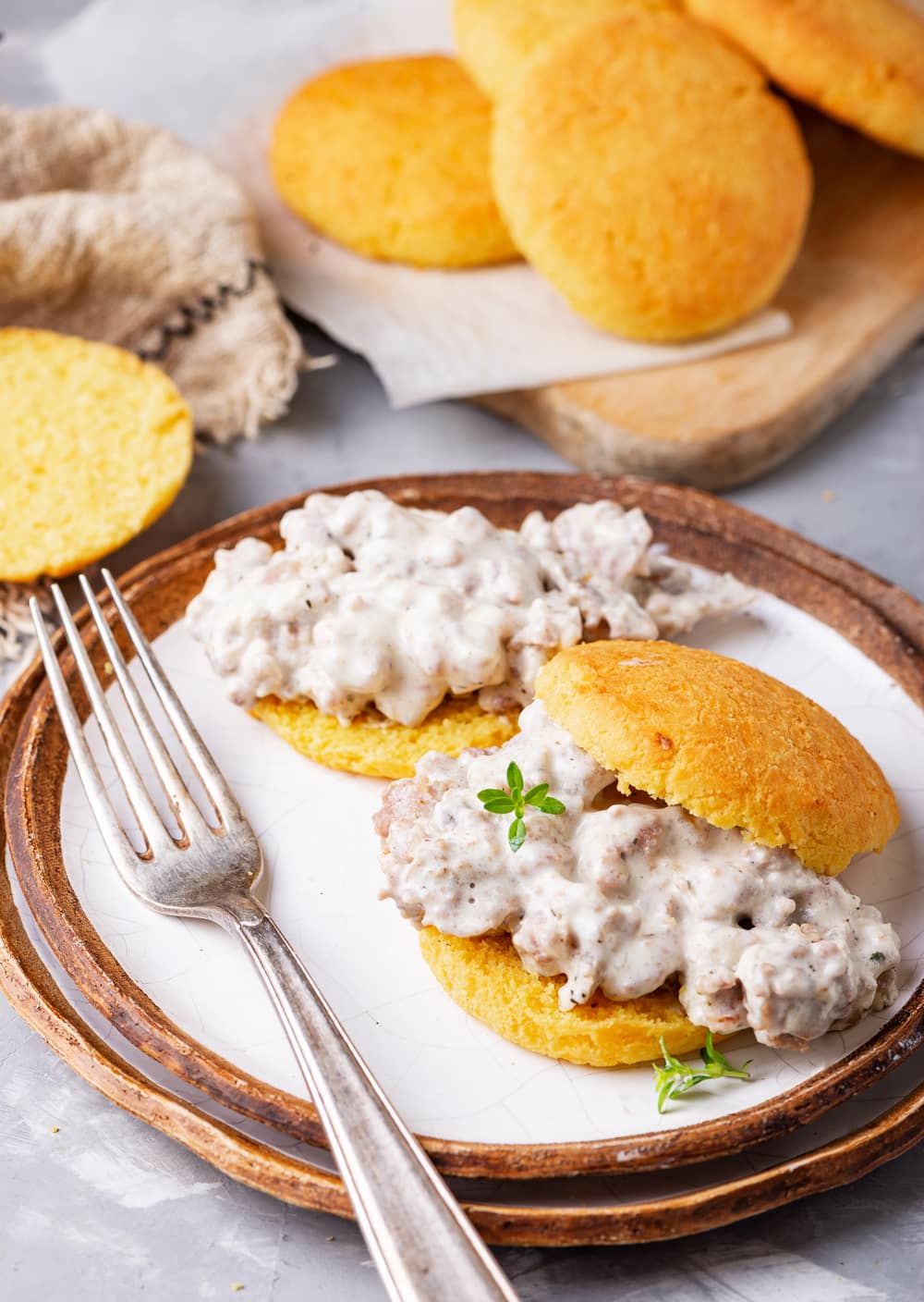 A biscuit filled with gravy and another biscuit behind it that's been topped with gravy. The biscuits are set on 2 plates that have been stacked on top of one another, and there are more biscuits on a cutting board behind the plate.