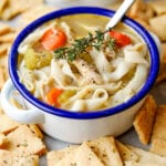 Chicken noodle soup in a white bowl with a blue rim. There are cheese crackers around the bowl, and a spoon in it.