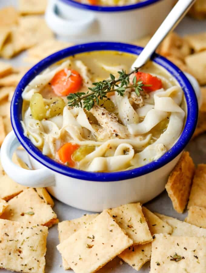 Chicken noodle soup in a white bowl with a blue rim. There are cheese crackers around the bowl, and a spoon in it.