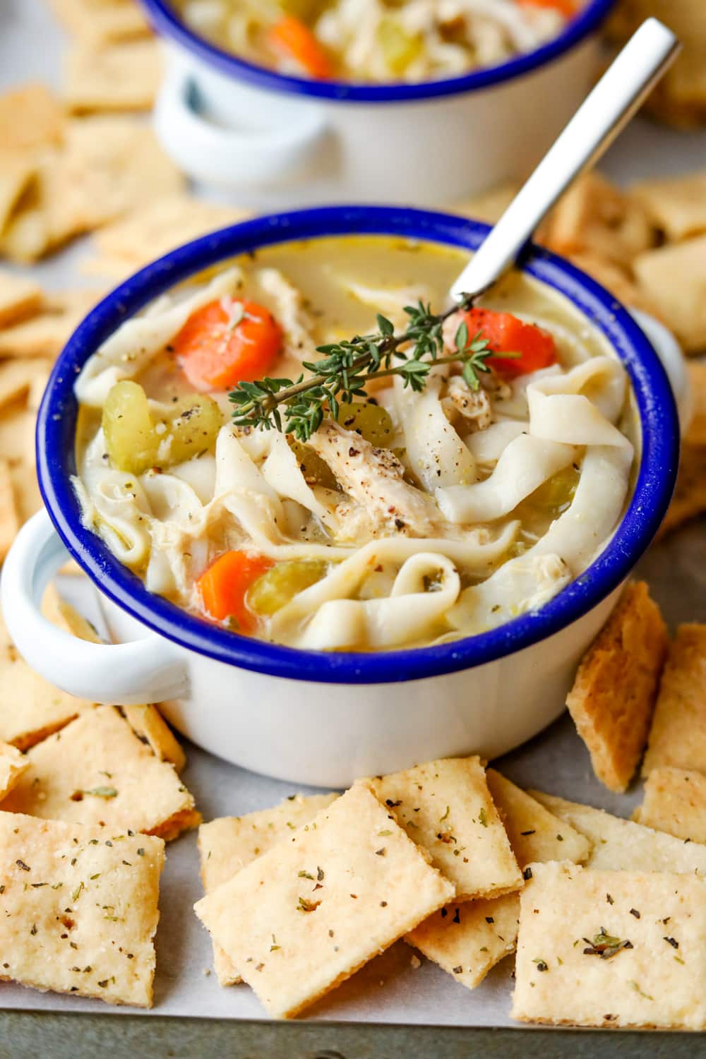 Chicken noodle soup in a white bowl with a blue rim. There are cheese crackers around the bowl, and a spoon in it.