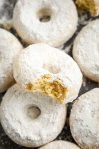 A powdered sugar donut with a bite taken out of it. The donut is stacked on top of other donuts.