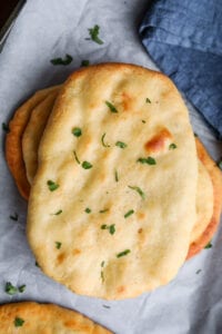 Naan breads stacked on top of each other. The naan is set on a white sheet of parchment paper, and a blue napkin is set next to it.