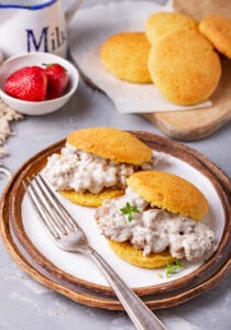 2 Biscuits on a plate that have been filled with gravy. There is a cutting board with more biscuits behind them, and a small bowl of strawberries next to the cutting board.