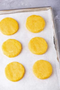 The dough of 6 biscuits that have been shaped, and placed on a baking sheet lined with white parchment paper.