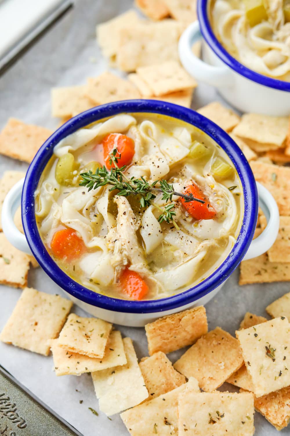 A bowl of chicken noodle soup set on a baking tray. There are crackers surrounding the soup.