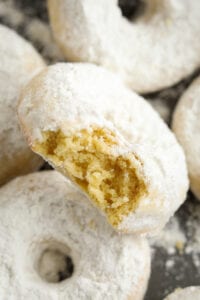 A donut with a bite taken out of it. The donut is covered in powdered sugar, and it's placed on top of other donuts.