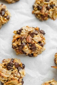 A cookie made with coconut flakes, pecans, and chocolate chips on parchment paper.