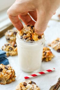 A hand dipping a cookie into a glass of milk. The glass is set on a tray of cookies, and there is a red and white stripped straw next to it.