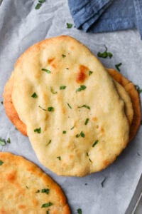 Naan bread stacked on top of one another on a sheet of parchment paper. The naan is topped with chopped up parsley.