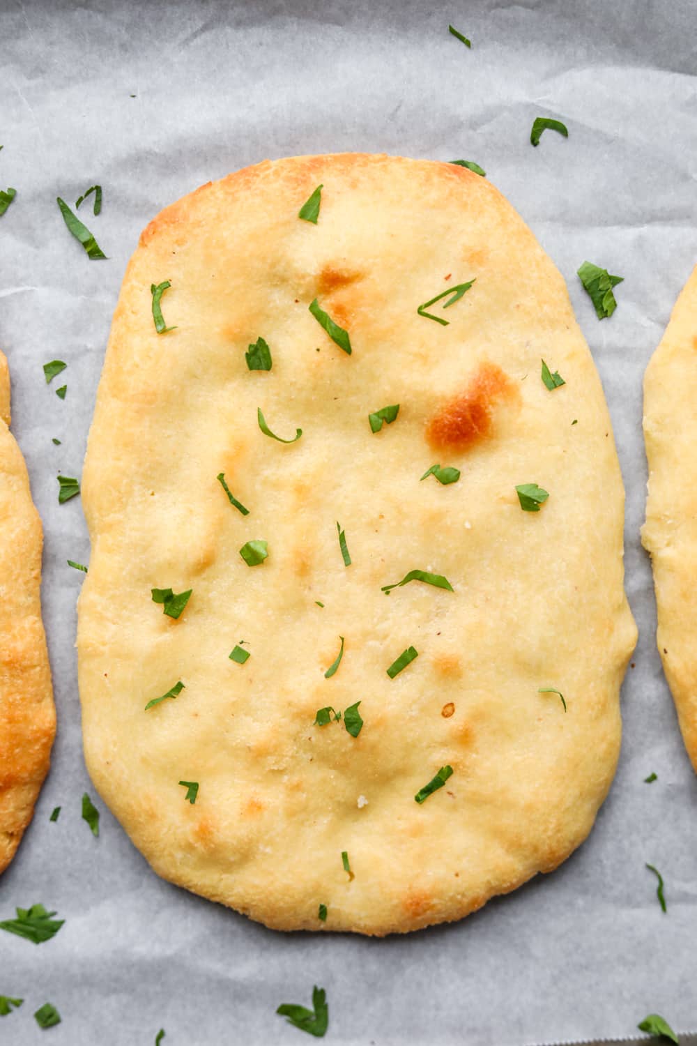 Naan bread with chopped parsley on top of it. The naan is set on a white sheet of parchment paper.