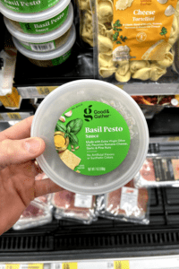 A hand holding a container of basil pesto sauce.