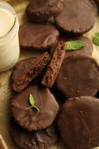 A chocolate coated cookie that's been broken in half and stacked on top of other chocolate cookies. There are mental leaves next to the cookies, and a glass of milk as well.