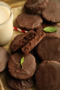 A chocolate cookie broken in half and stacked on other chocolate cookies. There's a glass of milk, a straw, and mint leaves next to the cookies.