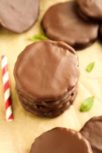 Chocolate cookies stacked on top of each other with mint leaves and more cookies next to them. There's also a red and white straw next to the cookies as well.