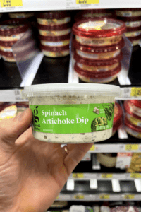 A hand holding a container of spinach and artichoke dip.