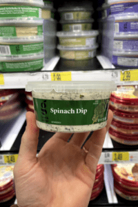 A hand holding a container of spinach dip.