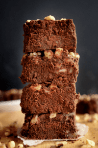 A stack of four brownies with walnuts on top of each one. There are a few walnuts on the wooden table the brownies are on.