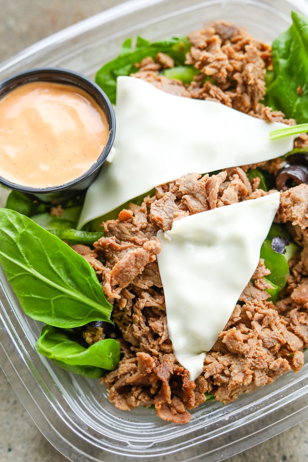 A plastic container filled with green vegetables, black olives, chopped up steak, two slices of white mozzarella cheese, and an orange dipping sauce.