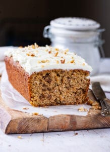 A loaf of carrot cake topped with a cream cheese frosting set on a cutting board lined with parchment paper.