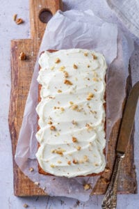 A loaf of carrot bread that's been topped with cream cheese frosting and crushed walnuts set on a cutting board.
