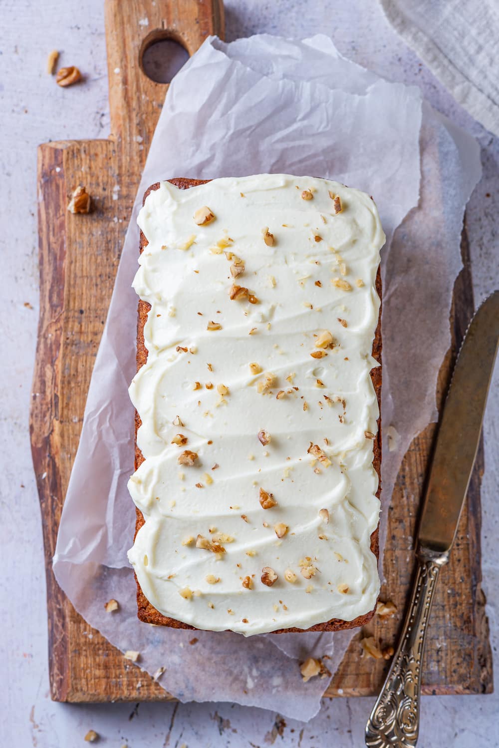 A loaf of carrot bread that's been topped with cream cheese frosting and crushed walnuts set on a cutting board.