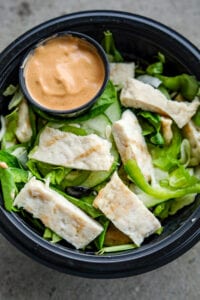 A black bowl filled with vegetables, a cup of orange dipping sauce, and cut up chicken.