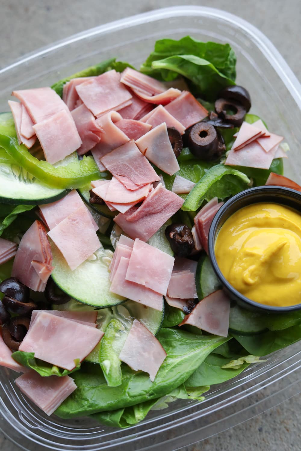 A plastic container filled with green vegetables, black olives, chopped up ham, and a cup of yellow mustard.