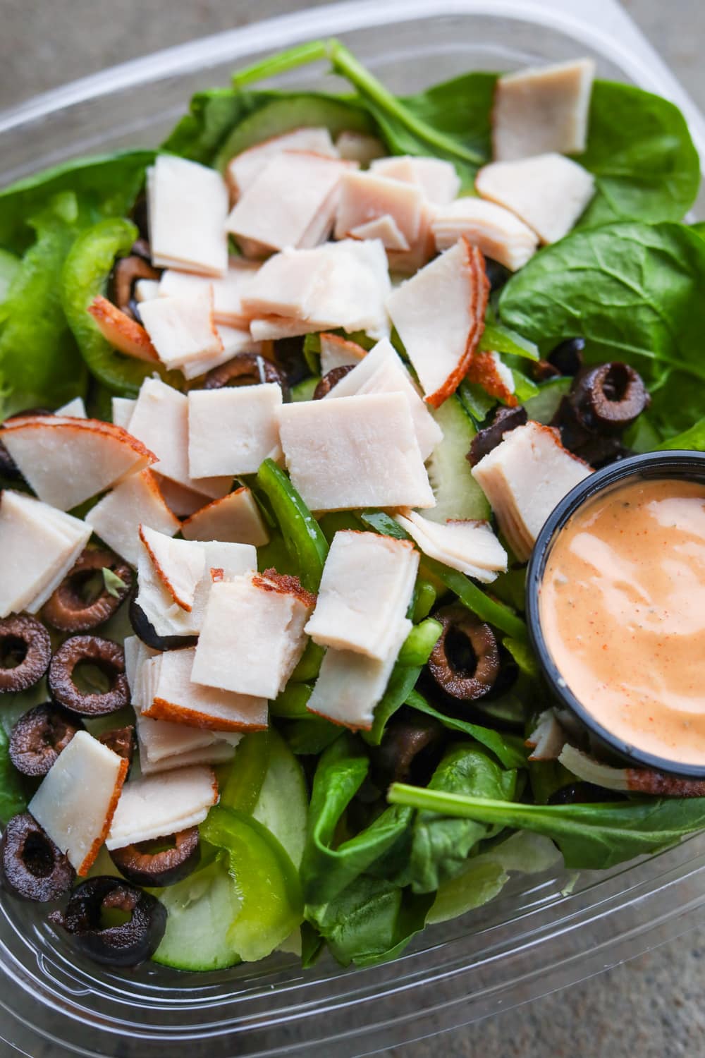 A plastic container filled with green vegetables, black olives, chopped up slices of turkey, and an orange dipping sauce.