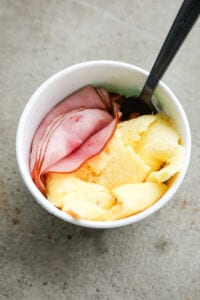 A white cup filled with slices of ham, scrambled eggs, and cheese.