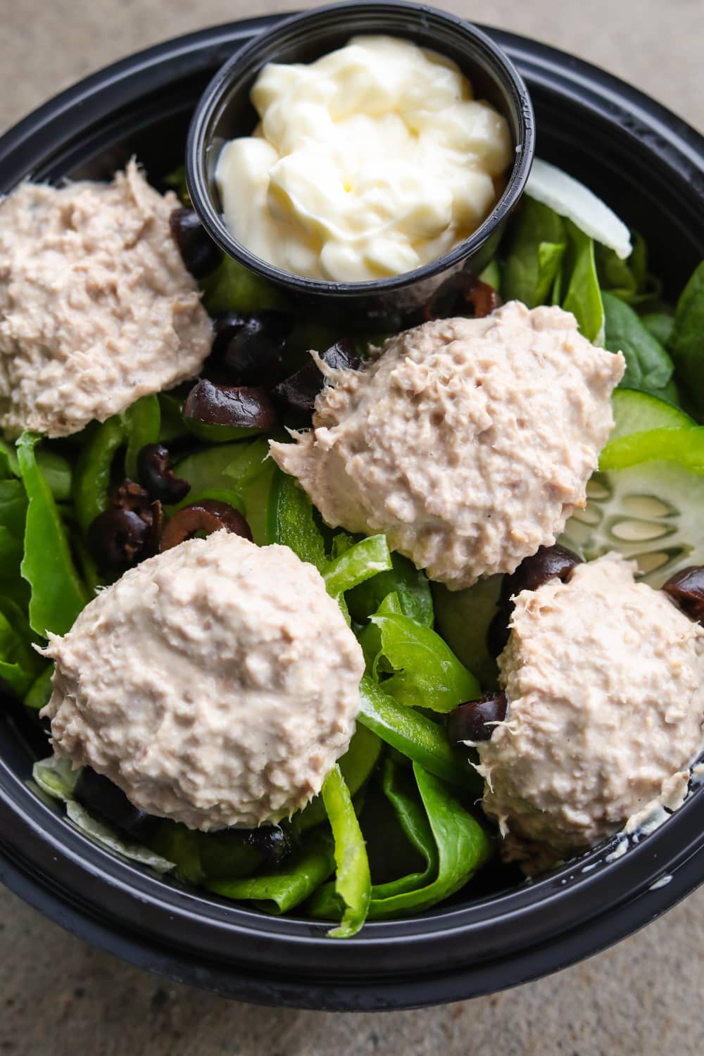 A black bowl filled with green vegetables, four scoops of tunafish that's mixed with mayonnaise, and a cup of mayonnaise.