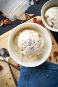 Ice cream in a white bowl set on a tan cutting board. There are silver spoons and a blue napkin next to the ice cream.